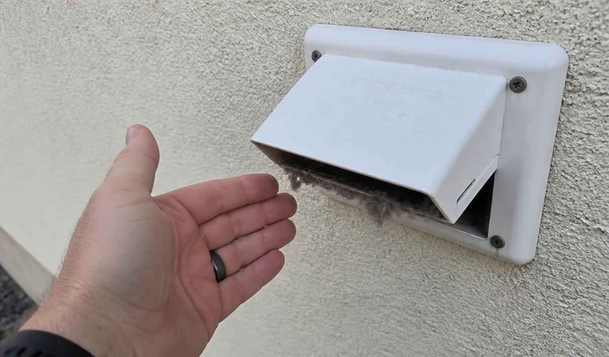 Homeowner checking airflow at an exterior dryer vent cap with lint around the flap on a Denair home.