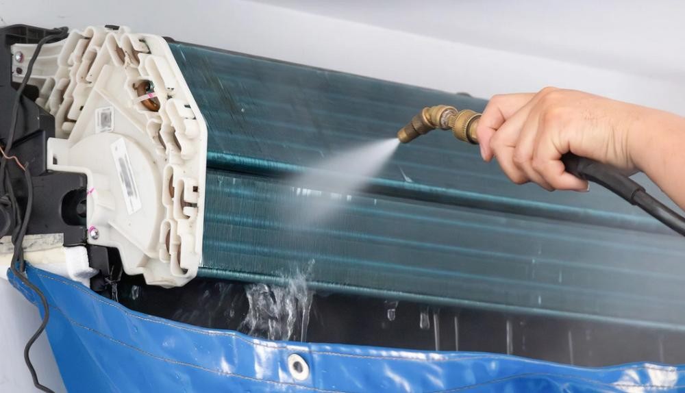 Technician cleaning an indoor AC evaporator coil with a pressurized water spray.